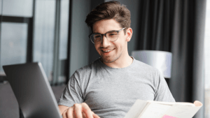 Hombre joven sonriente con gafas utilizando su ordenador portátil en casa, buscando información sobre cómo elegir una compañía eléctrica fiable y con tarifas de luz transparentes